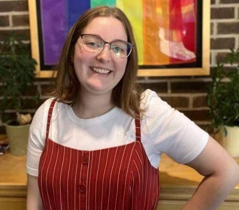 Person smiling in front of hanging LGBTQ+ pride flag.